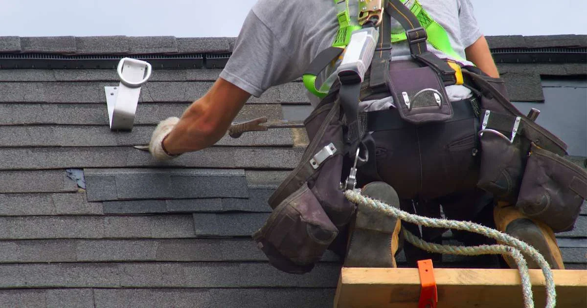 A roofing contractor in safety gear installing or repairing asphalt shingles on a sloped roof.